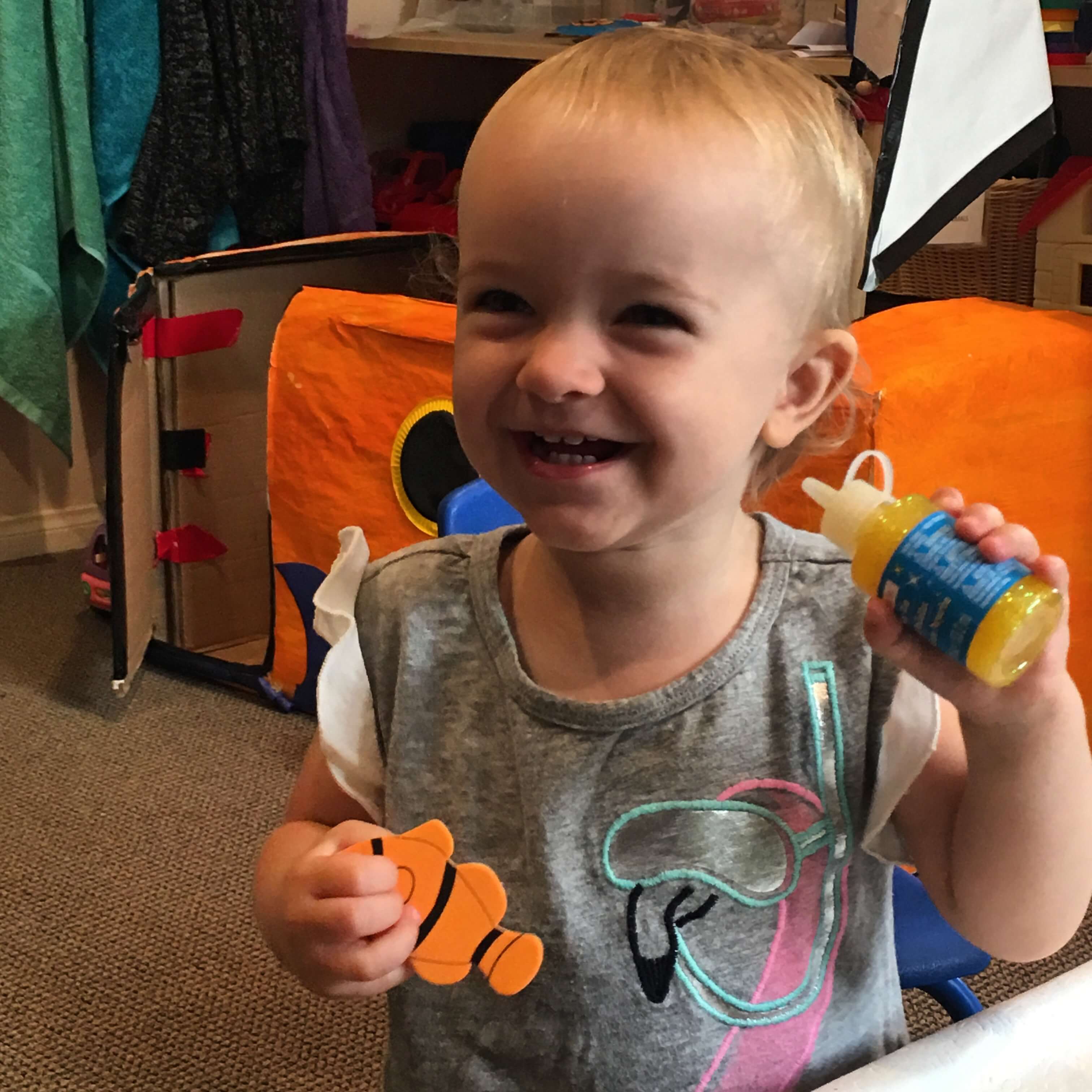 toddler smiling wiht a sticker of a fish and glue to decorate her boat at Early Childhood Development Associates' class