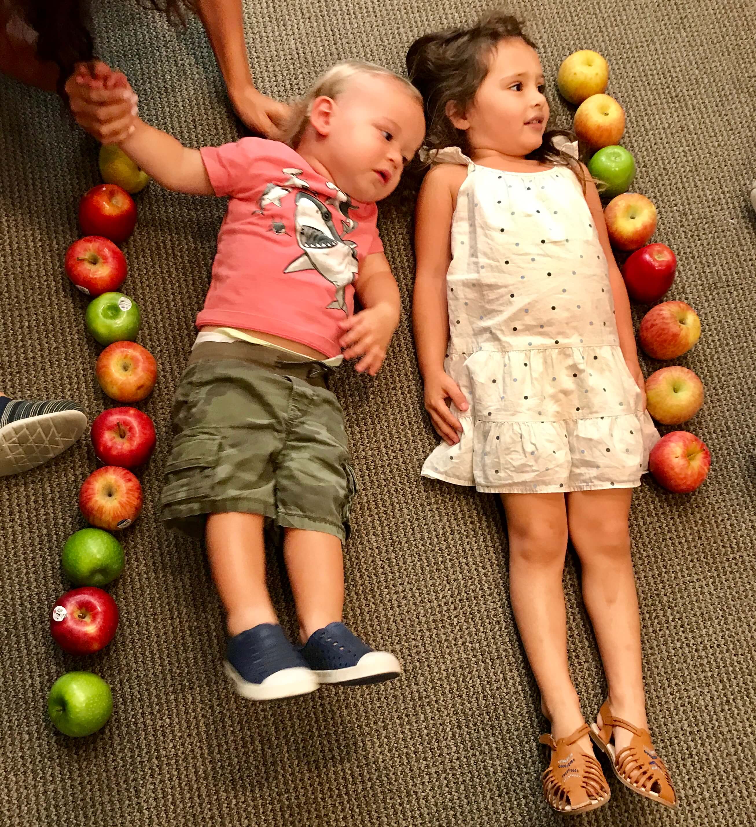 2 toddlers luong on the floor with apples beside them in the form of a line to measure their height with the apples at ECDA